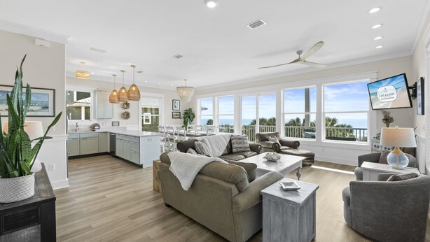 A spacious living area with a large sofa, a coffee table, and a TV mounted on the wall. There is a balcony with a view of the ocean.