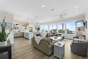 A spacious living area with a large sofa, a coffee table, and a TV mounted on the wall. There is a balcony with a view of the ocean.