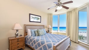 A bedroom with a bed, nightstands, a ceiling fan, a large window with a view of the beach, and a framed fish picture on the wall.