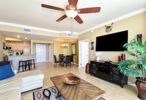 A spacious living room with a ceiling fan, a dining area, a kitchen, and a large TV.