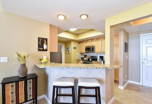 A kitchen with a granite countertop, two bar stools, wooden cabinets, a refrigerator, a microwave, and a vase with dried plants.