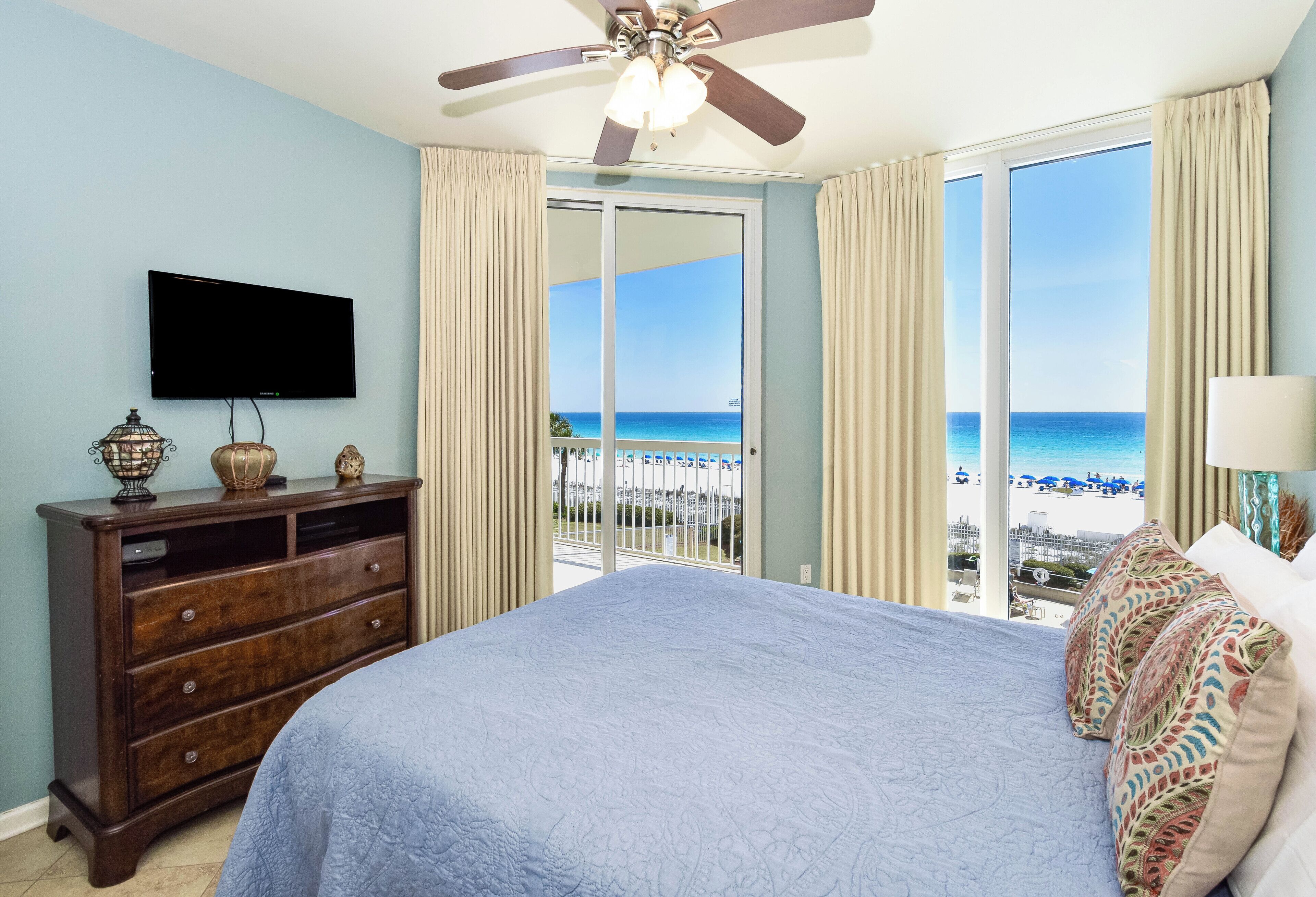 A bedroom with a bed, a TV, a dresser, and a view of the beach.