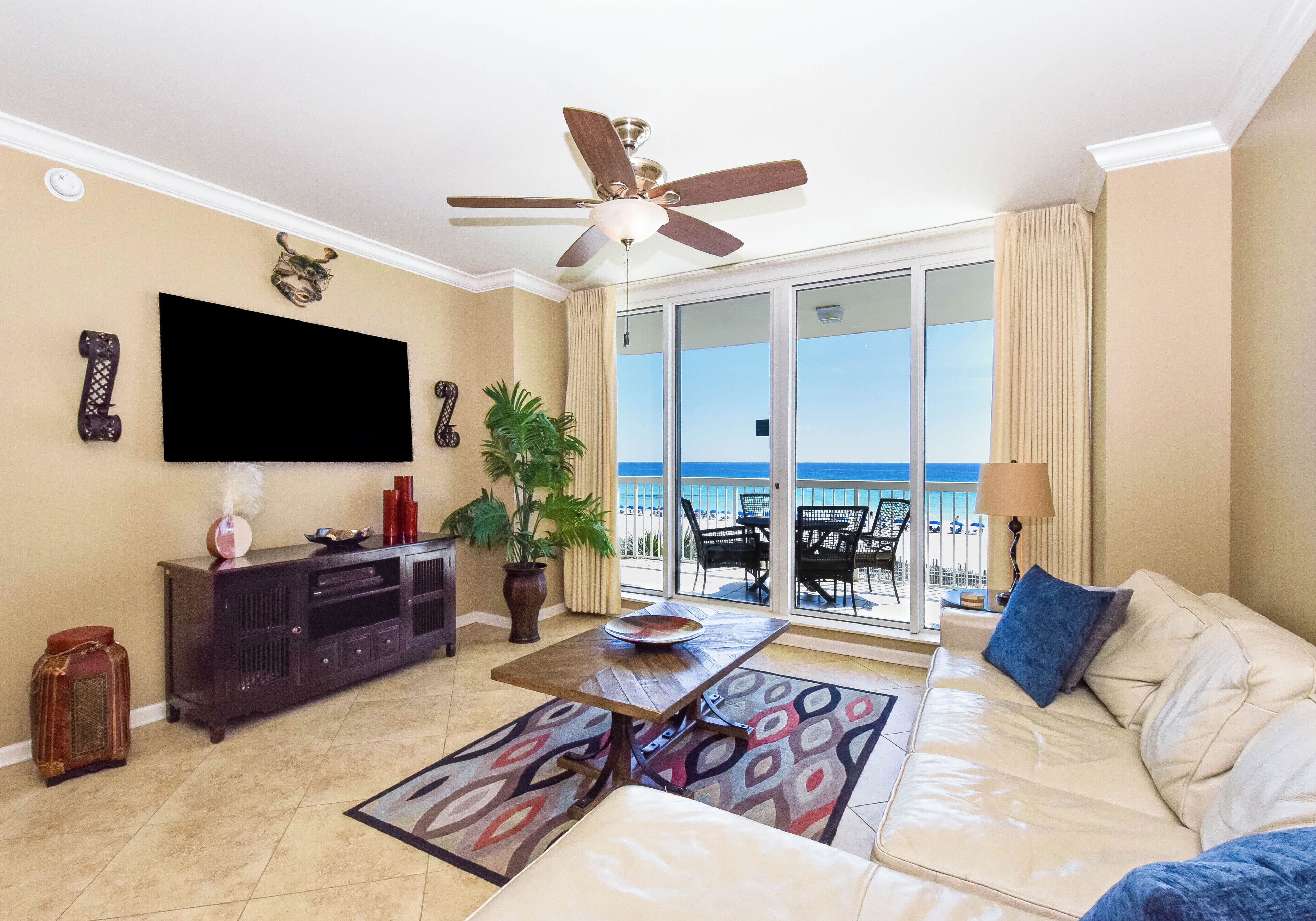 A living room with a sofa, a coffee table, a TV, a ceiling fan, a potted plant, and a view of the beach through large windows.