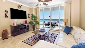 A living room with a sofa, a coffee table, a TV, a ceiling fan, a potted plant, and a view of the beach through large windows.