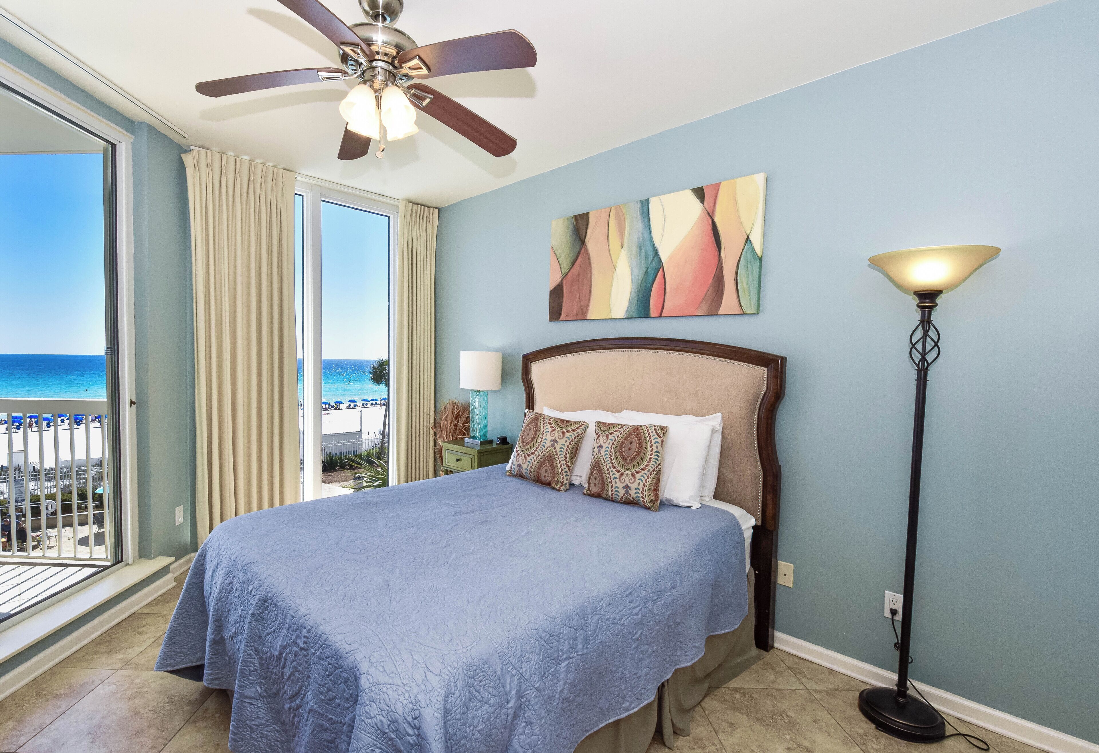 Une chambre d’hôtel avec un lit, un ventilateur de plafond, une table de chevet avec une lampe, un tableau au mur et une vue sur la plage.