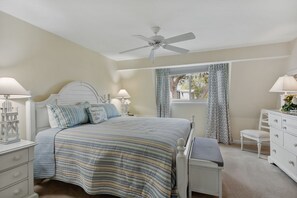 Un dormitorio con cama, mesitas de noche, cómoda, silla, ventilador de techo y ventana con cortinas.