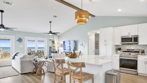 A modern kitchen with white cabinets, a stainless steel microwave, and an oven. There is a dining area with wooden chairs and a table, and a large window overlooking the ocean. - Beachfront | Pet Friendly | Renovated Coastal Cottage (Gulf Shores)