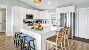A modern kitchen with white cabinets, stainless steel appliances, a large island with stools, and a wicker pendant light. - Beachfront | Pet Friendly | Renovated Coastal Cottage (Gulf Shores)