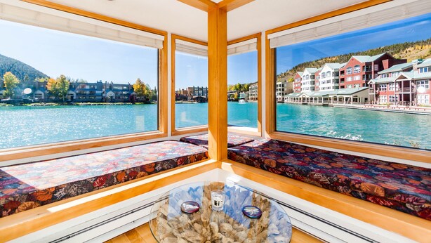 A room with large windows offering views of a lake and buildings. There are cushioned seating areas with patterned upholstery.