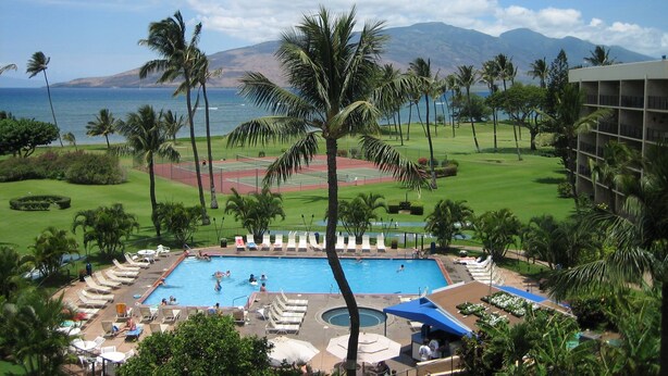 A hotel pool area with a tennis court, lounge chairs, and a view of the ocean and mountains. - Maui Sunset Vacation Rentals B514 (Kihei)
