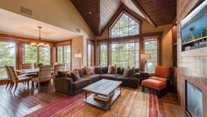 A spacious living area with a large wooden coffee table, a sofa, and a fireplace. There are multiple windows providing natural light and views of the outdoors.