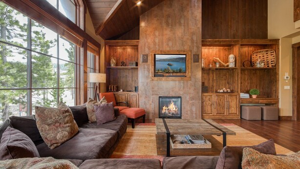 A spacious living room with a large fireplace, wooden furniture, and a view of the outdoors through large windows.