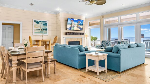 A spacious living room with a blue sofa set, a dining table with chairs, a fireplace, and a large TV mounted on the wall. - CASA DEL SOL - 4BR 4 BTH BEACHFRONT HOME W/POOL located in The Plantation (St, George Island)