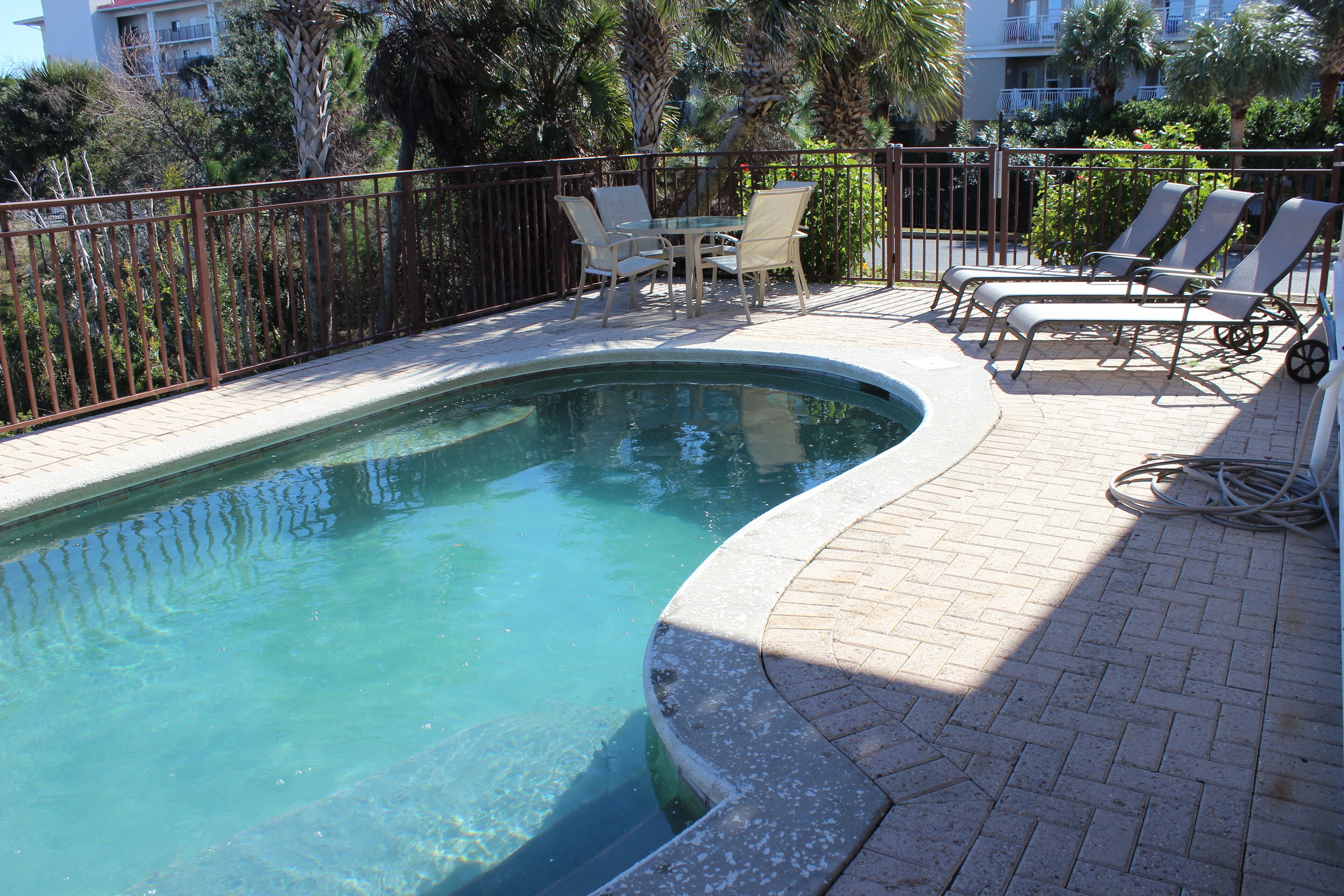 A swimming pool with a curved edge, surrounded by a brick patio. There are lounge chairs and a table with chairs.