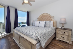 A bedroom with a large bed, a nightstand, a ceiling fan, and a view of the outdoors.
