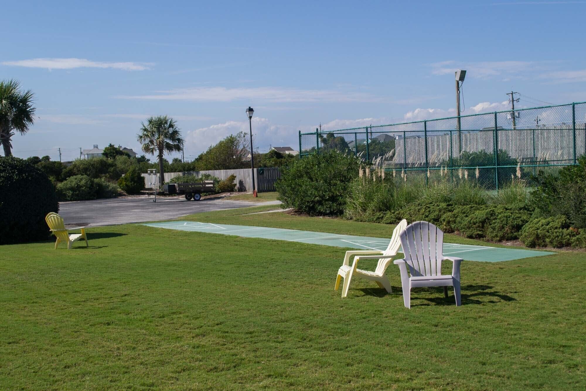 A grassy area with two Adirondack chairs, a green tennis court, and a chain-link fence.