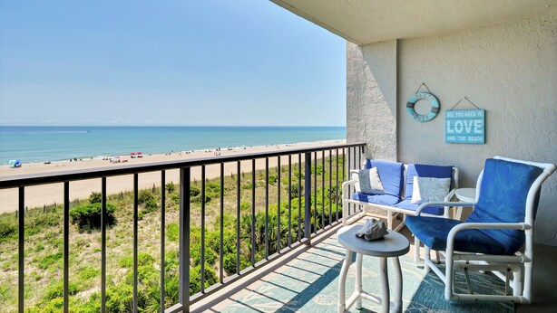 Un balcon avec vue sur la plage, deux chaises bleues, une petite table et deux éléments de décoration murale.