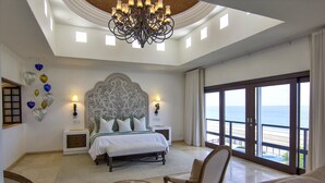 A spacious bedroom with a large bed, a chandelier, and a view of the beach through large windows.