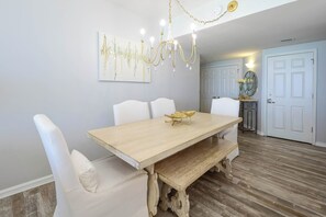 A dining area with a wooden table, white upholstered chairs, a chandelier, and a wall-mounted abstract painting. - Refreshing coastal beachfront condo w/updated flooring! (Destin)