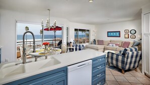 A modern kitchen with a view of the beach, a dining area with a red umbrella, and a living room with a sofa and decorative pillows. - Panoramic Ocean View White Sandy Beaches   Community Jacuzzi (Oceanside)