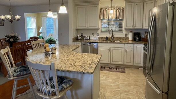 A kitchen with white cabinets, a granite countertop, a stainless steel refrigerator, and a dining area with wooden chairs and a table. - Chic Gem in The Villages - Modern Amenities & Prime Location! Call today! (The Villages)