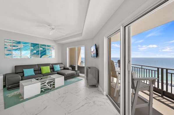A modern living room with a sofa, coffee table, and artwork, leading to a balcony with a view of the ocean.