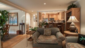 A living room with a sofa, a coffee table, and a lamp. There is a kitchen in the background with wooden cabinets and a microwave. - Ocean Pines #28 - Pebble Beach Condo - Oceanview Luxury (Pebble Beach)
