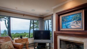 A living room with a television, a fireplace, and a painting of a beach scene. - Ocean Pines #28 - Pebble Beach Condo - Oceanview Luxury (Pebble Beach)