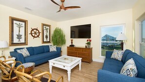 A living room with a blue sofa, wicker chairs, a white coffee table, a wooden cabinet, and a ceiling fan. - Grace | 6BR Fort Morgan Beach Retreat | Pet-Friendly | Gulf View (Gulf Shores)