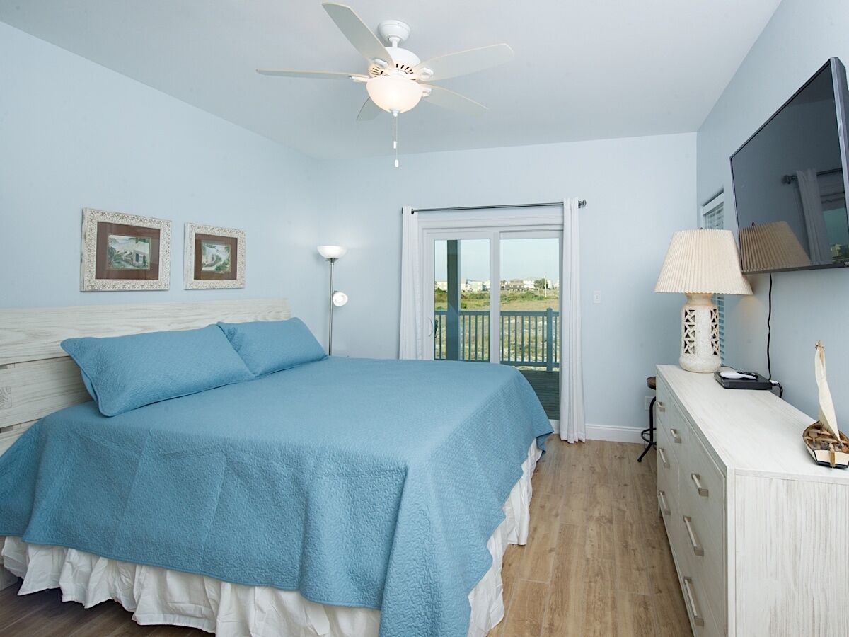 A bedroom with a bed, a ceiling fan, a dresser, a lamp, and a view of the outdoors.