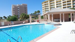 A swimming pool with a sunshade structure, surrounded by a white tiled deck and a building with multiple balconies. - Grace | 6BR Fort Morgan Beach Retreat | Pet-Friendly | Gulf View (Gulf Shores)