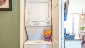 Una lavadora de carga frontal con detergente Tide, un comedor con sillas y una mesa, y un televisor montado en la pared.