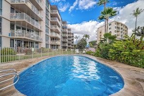 A swimming pool surrounded by multi-story buildings with balconies, palm trees, and a clear sky.