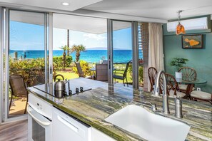 A kitchen with a view of the ocean, a dining area with chairs, and a table. - Kamaole Nalu 106-Unbeatable Proximity to the Shore & Unbelievable Locale Await (Kihei, Maui)