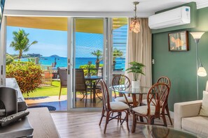 A room with a glass door leading to a balcony with a view of the sea. There is a table with chairs, a sofa, and a TV. - Kamaole Nalu 106-Unbeatable Proximity to the Shore & Unbelievable Locale Await (Kihei, Maui)