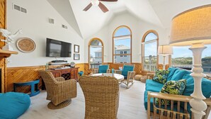 A spacious living room with a high ceiling, large windows, wicker furniture, and a television. - Time To Coast: Semi-Oceanfront, Hot Tub, Ocean Views (Corolla)