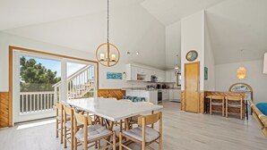 A spacious kitchen with white cabinets, a dining area with a white table and wooden chairs, and a large window with a view of trees. - Time To Coast: Semi-Oceanfront, Hot Tub, Ocean Views (Corolla)