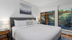 Ein Schlafzimmer mit einem großen Bett, einem Nachttisch mit Lampe, einem Fenster mit Blick auf Bäume und einem Gemälde an der Wand.
