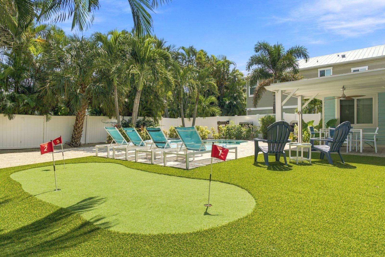 Zona de piscina con sillones de descanso y una piscina pequeña, rodeada de palmeras y una cerca blanca.