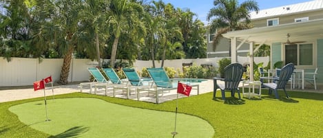 Zona de piscina con sillones de descanso y una piscina pequeña, rodeada de palmeras y una cerca blanca.