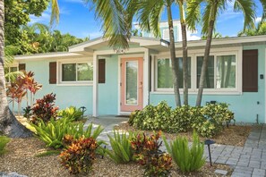 A modern house with a light blue exterior, a front door, and a well-maintained garden with various plants and a palm tree. - Ebb Tide by Duncan Real Estate (Holmes Beach)