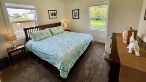 Un dormitorio con una cama, dos mesitas de noche, una lámpara y un aparador. Hay dos ventanas con vistas a zonas verdes y una cerca de piquetes blancos.