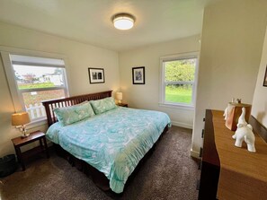 A bedroom with a bed, two bedside tables, a lamp, and a dresser. There are two windows with views of greenery and a white picket fence. - Potter's Bungalow - Near Downtown Plaza, 3 bed (Arcata)