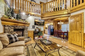 A spacious living room with a stone fireplace, a wooden dining area, and a kitchen with wooden cabinets and a large clock. - Downtown Cabin w/Pool Table|Pool Access (PIGEON FORGE)