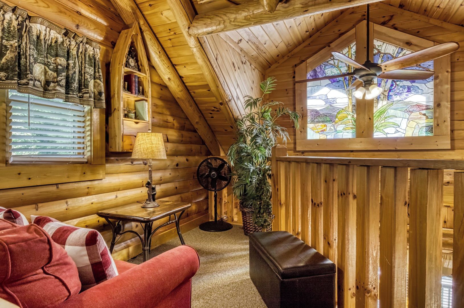 A cozy log cabin interior with a red sofa, a table with a lamp, a fan, and a large window with a view of greenery.