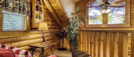 A cozy log cabin interior with a red sofa, a table with a lamp, a fan, and a large window with a view of greenery.