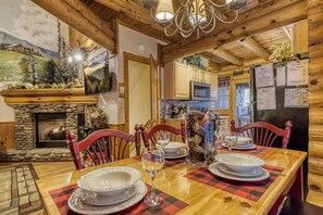 Interior de madera con chimenea de piedra, mesa de comedor para cuatro personas y una cocina visible al fondo.