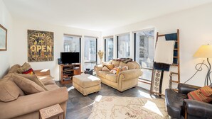 Ein geräumiges Wohnzimmer mit einem beigefarbenen Sofa, einem schwarzen Ledersessel und einem Couchtisch. Ein Fernseher steht auf einem Holzgestell, eine Leiter lehnt an der Wand und ein großes Fenster lässt natürliches Licht herein.
