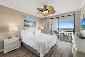 A hotel room with a bed, nightstands, a ceiling fan, a painting, a balcony with a view of the beach, and a dining area.