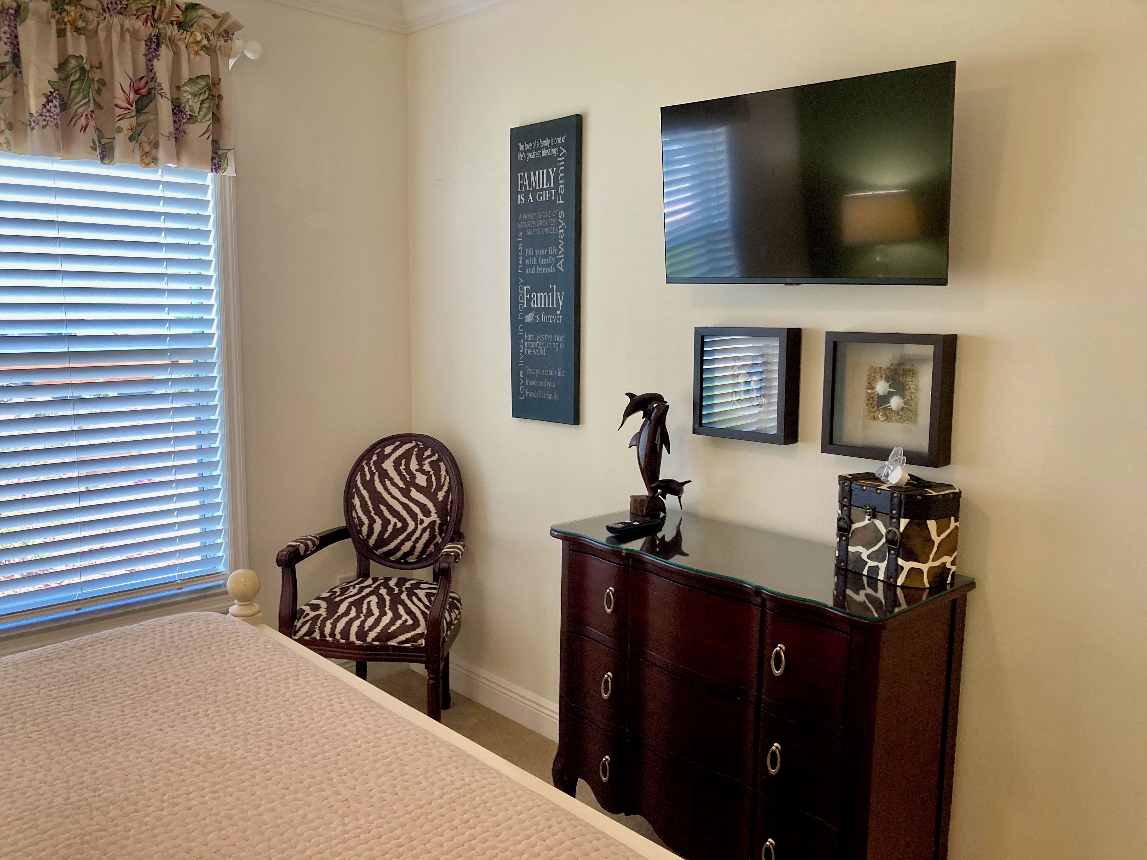 Un dormitorio con cama, una silla, cómoda, televisor y adornos en la pared.